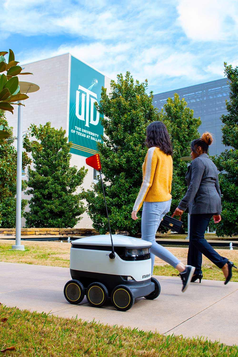 A Starship delivery robot passes by two women as it travels through Margaret McDermott Mall on the UT Dallas campus. The machine is roughly the size and shape of a cooler on six wheels, with an antenna topped by a reflector, and band of sensors and lights around its top edge.