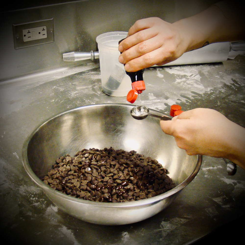 A chef pouring a clear liquid into a measuring spoon over a steel bowl full of chocolate chips.