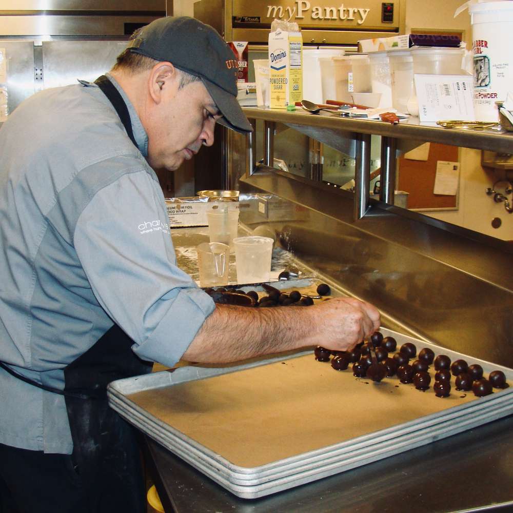 A chef spooning a chocolate-covered cake ball onto a tray filled with more such pastries.